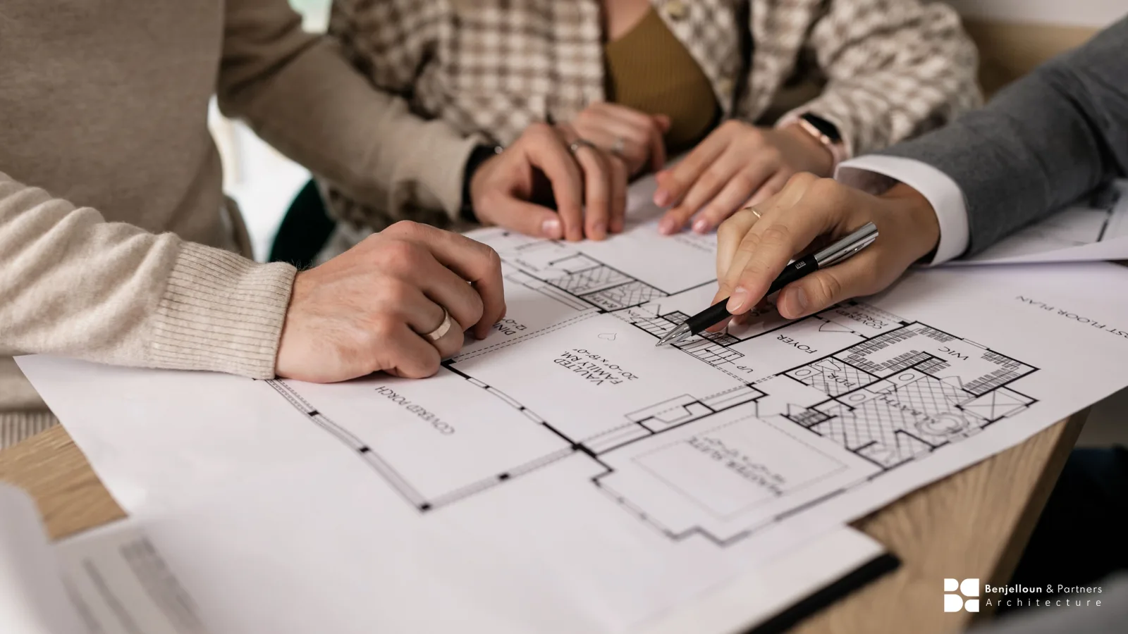 Three people gathered around a table reviewing a printed architectural floor plan, one indicating a detail with a pen.